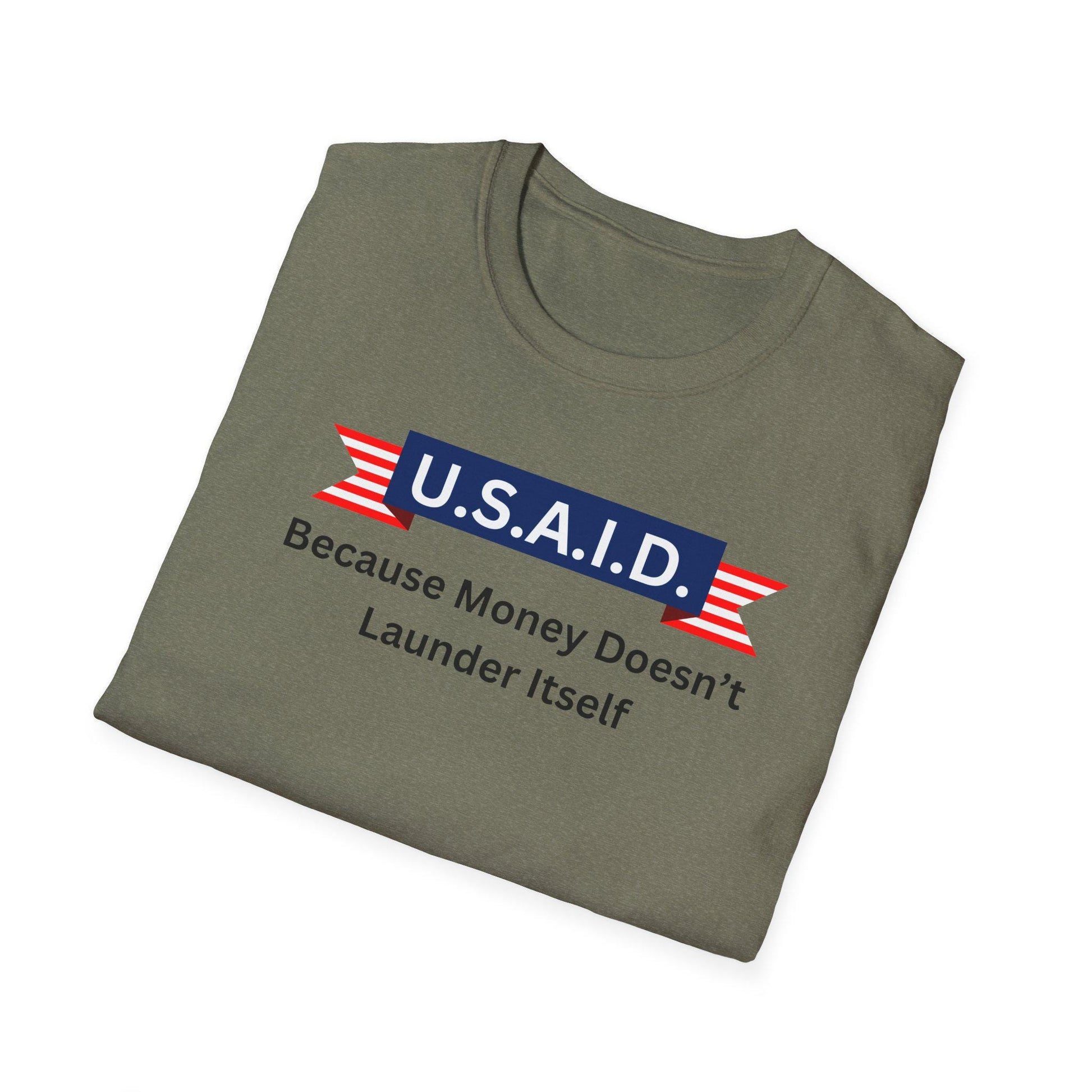 A military green T-shirt is neatly folded against a white background. The shirt features a bold, satirical graphic on the front. A navy blue banner with white letters reads &quot;U.S.A.I.D.&quot; and is accented with red and white striped ribbon elements. Below the banner, black text reads: &quot;Because Money Doesn’t Launder Itself.&quot; The design has a humorous political message with a patriotic visual style.