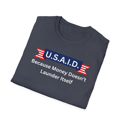 A folded navy blue T-shirt is displayed on a white background. The shirt features a bold satirical design with a navy blue banner labeled &quot;U.S.A.I.D.&quot; in white letters, accented by red and white striped ribbon graphics on either side. Below the banner, the white text reads: &quot;Because Money Doesn’t Launder Itself.&quot; The design mimics a government agency logo with a humorous political commentary.