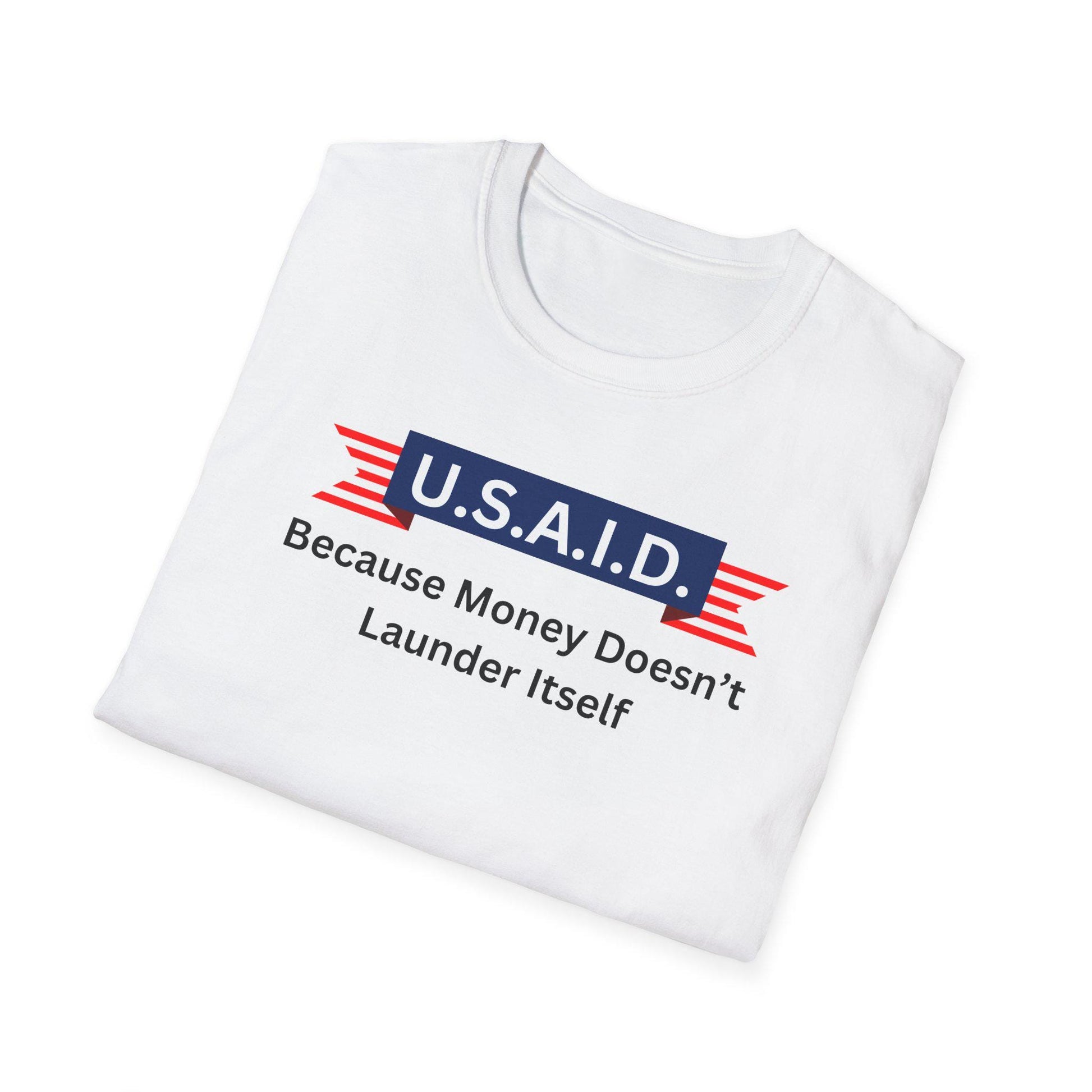 A white T-shirt is neatly folded on a white background. The shirt features a bold satirical graphic with a navy blue banner that reads &quot;U.S.A.I.D.&quot; in white letters, flanked by red and white striped ribbon graphics. Below the banner, black text reads: &quot;Because Money Doesn’t Launder Itself.&quot; The design mimics a government agency logo with a humorous political twist.