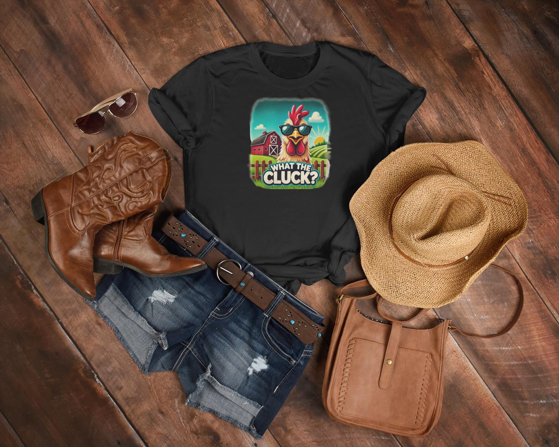 Flat lay of a country-inspired outfit featuring a black t-shirt with a colorful cartoon chicken wearing sunglasses and the phrase &quot;What the Cluck?&quot; set against a farm background. The ensemble includes distressed denim shorts, a brown leather belt, cowboy boots, a straw hat, a brown crossbody bag, and aviator sunglasses, all arranged on a rustic wooden floor.