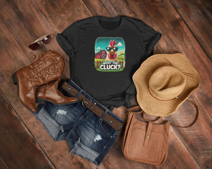 Flat lay of a country-inspired outfit featuring a black t-shirt with a colorful cartoon chicken wearing sunglasses and the phrase &quot;What the Cluck?&quot; set against a farm background. The ensemble includes distressed denim shorts, a brown leather belt, cowboy boots, a straw hat, a brown crossbody bag, and aviator sunglasses, all arranged on a rustic wooden floor.