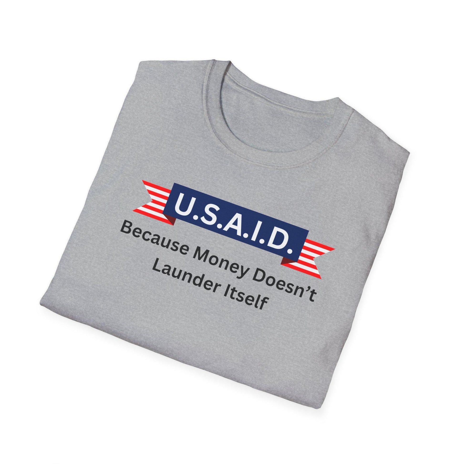 A light gray T-shirt is neatly folded on a white background. The shirt features a bold, satirical graphic with a navy blue banner labeled &quot;U.S.A.I.D.&quot; in white text, flanked by red and white striped ribbon designs. Beneath the banner, black text reads: &quot;Because Money Doesn’t Launder Itself.&quot; The design parodies a government agency logo with a humorous political message.