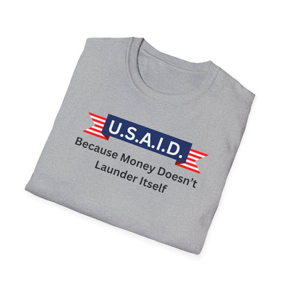 A light gray T-shirt is neatly folded on a white background. The shirt features a bold, satirical graphic with a navy blue banner labeled &quot;U.S.A.I.D.&quot; in white text, flanked by red and white striped ribbon designs. Beneath the banner, black text reads: &quot;Because Money Doesn’t Launder Itself.&quot; The design parodies a government agency logo with a humorous political message.