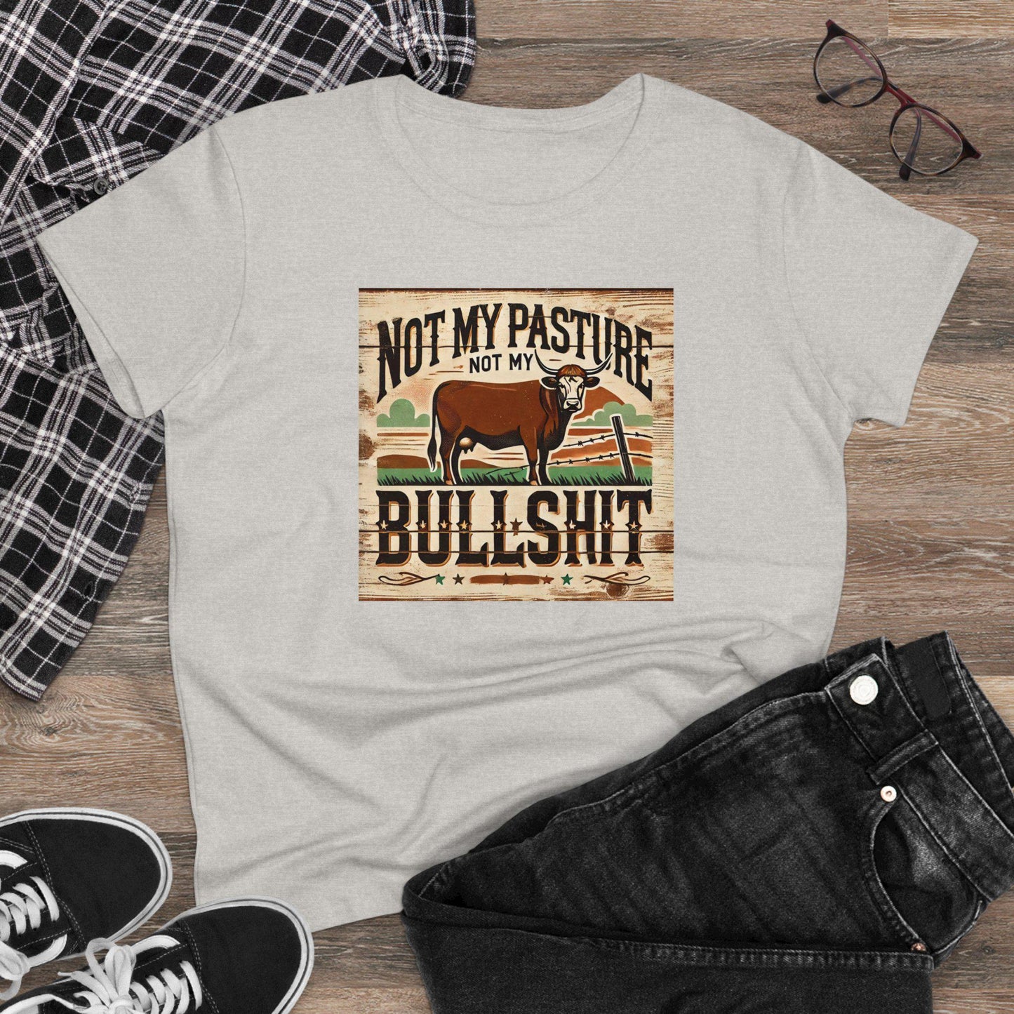 A light gray T-shirt is laid flat on a wooden surface, surrounded by casual accessories including a black-and-white plaid shirt, black jeans, black sneakers, and a pair of eyeglasses. The T-shirt features a rustic, vintage-style graphic with a brown cow standing in a pasture next to a broken fence. Bold, distressed text reads: &quot;Not my pasture, not my bullshit.&quot; The design evokes a country, no-nonsense attitude with earthy tones and a wood-textured background.