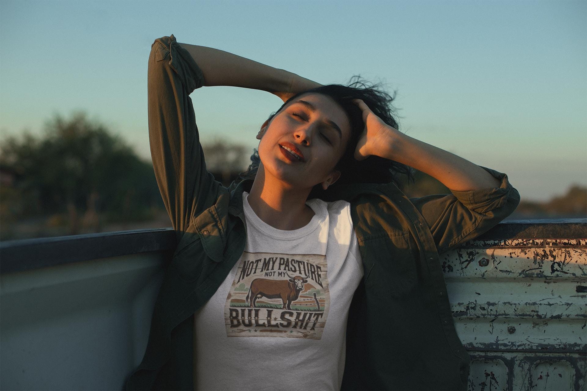 A woman relaxes in the back of a weathered pickup truck at sunset, with her hands behind her head and eyes closed. She wears an olive green button-up shirt over a white T-shirt that features a rustic graphic of a cow and the bold, humorous text: &quot;Not my pasture, not my bullshit.&quot; The scene conveys a laid-back, country-inspired vibe.