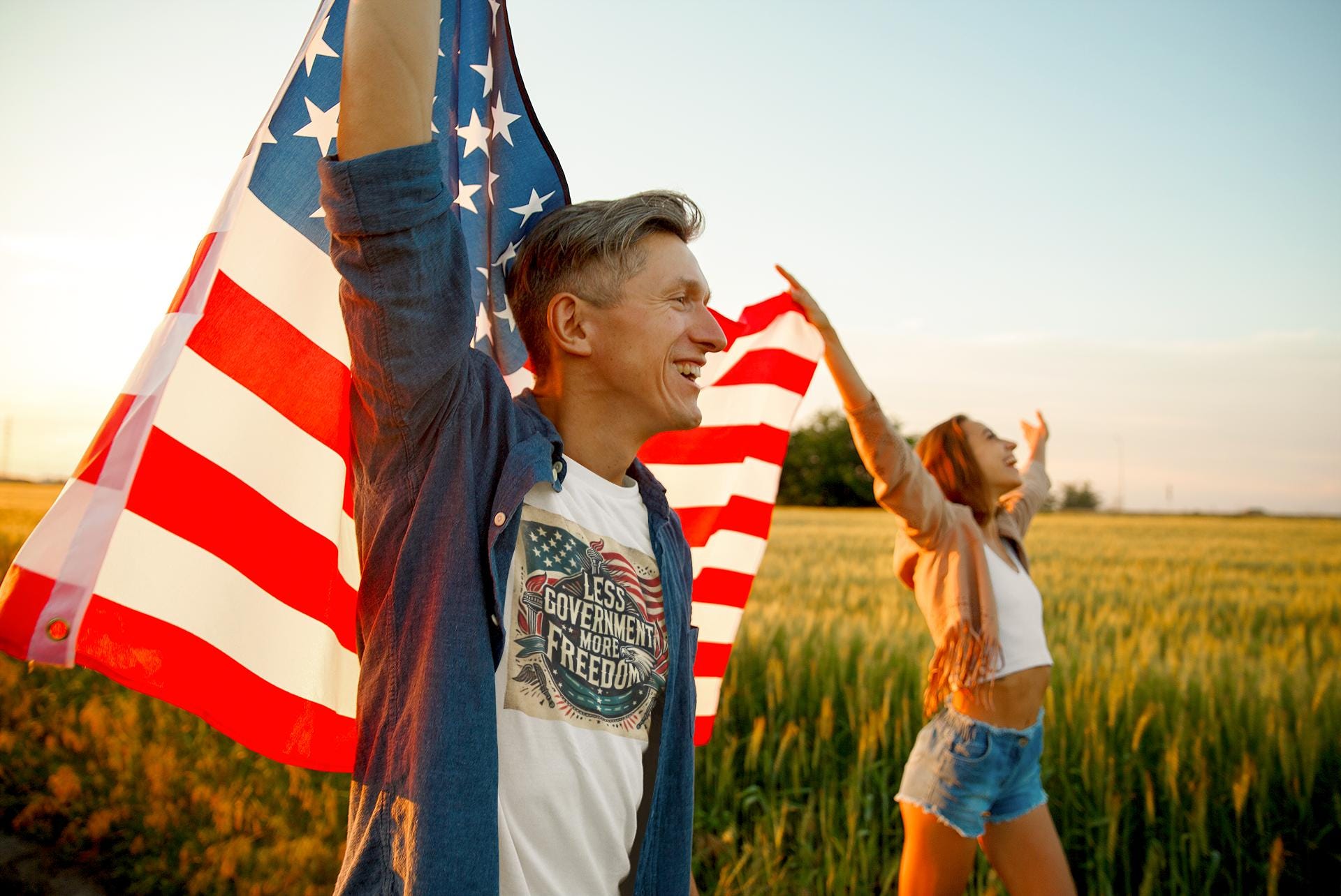 Smiling man and woman celebrating freedom outdoors with American flags, patriotic t-shirt slogan &#39;Less Government More Freedom&#39; during golden hour in a wheat field.