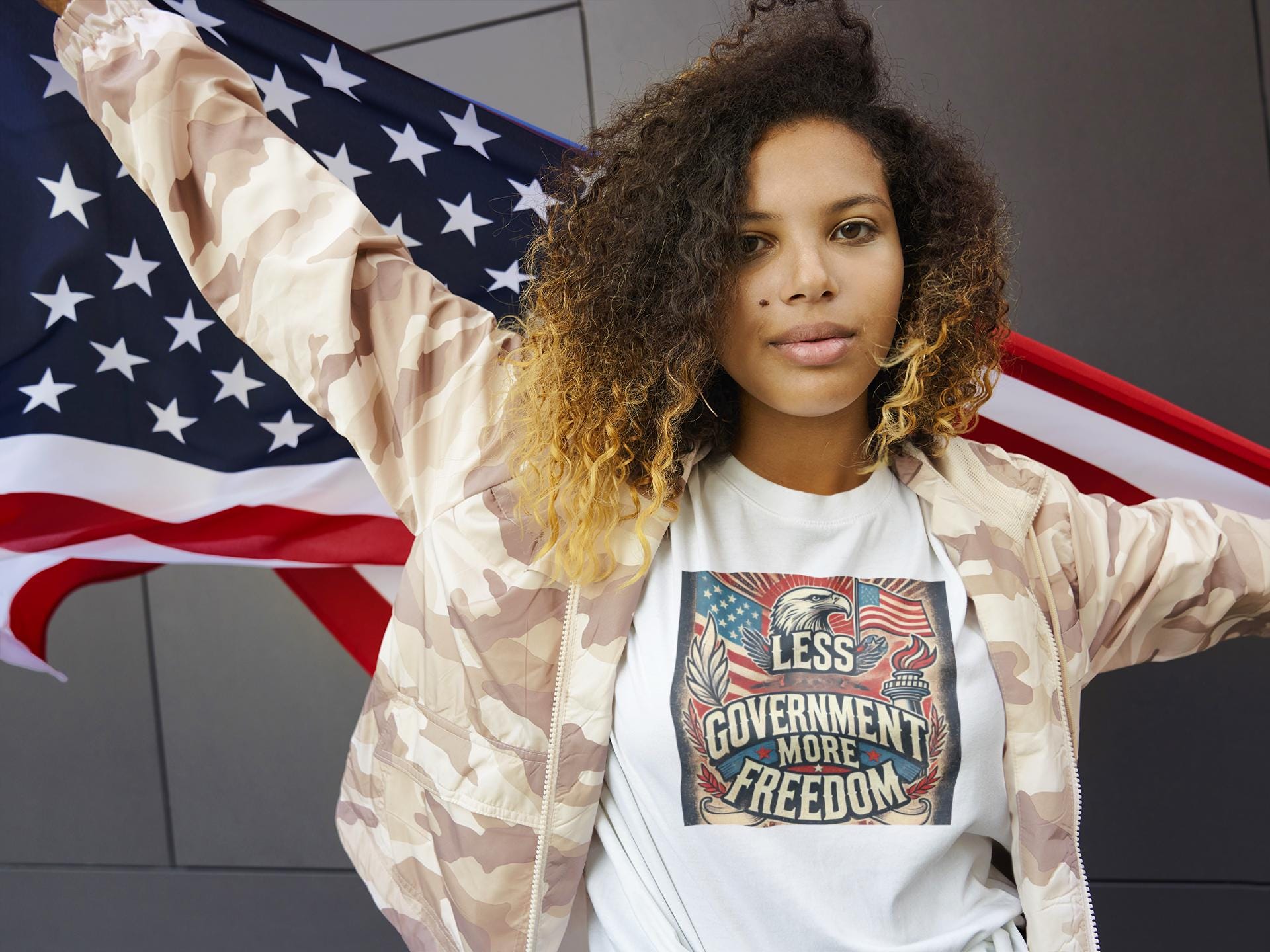 Confident woman wearing white &#39;Less Government More Freedom&#39; patriotic t-shirt and camouflage jacket while holding American flag — bold conservative fashion celebrating liberty and independence.
