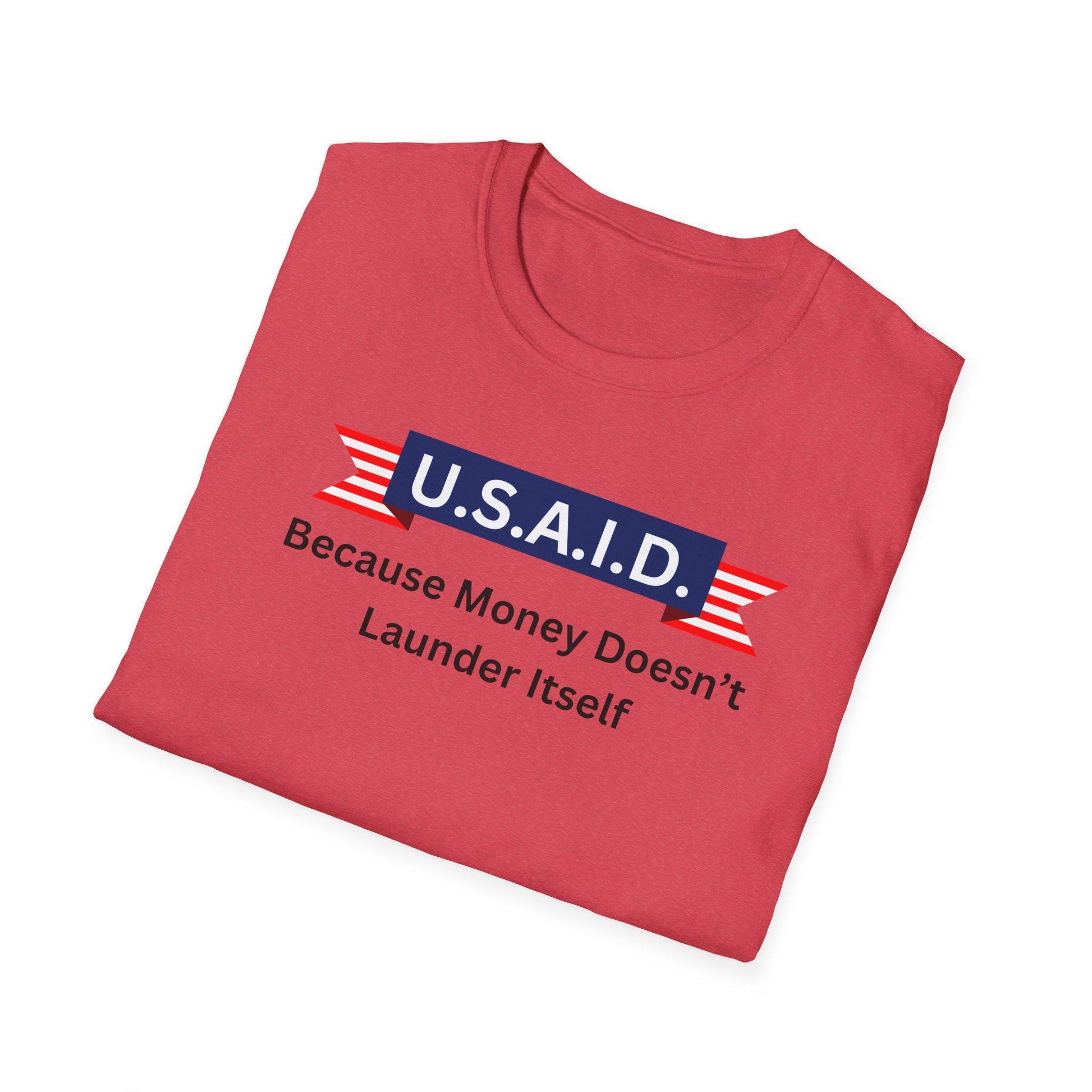 A red T-shirt is neatly folded against a white background. Printed on the front is a satirical design resembling a patriotic banner. It features a blue box with white letters reading &quot;U.S.A.I.D.&quot; flanked by red and white striped ribbons. Below, in black text, it says: &quot;Because Money Doesn’t Launder Itself.&quot; The design plays on political humor with a clean, bold layout.