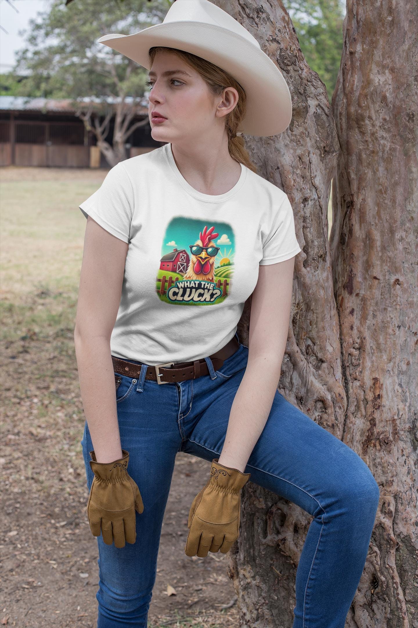Woman dressed in a western outfit, wearing a white graphic t-shirt with a cartoon chicken in sunglasses and the phrase &quot;What the Cluck?&quot; printed on it. She is also wearing a white cowboy hat, brown gloves, blue jeans, and a brown belt, leaning against a tree in an outdoor ranch setting.