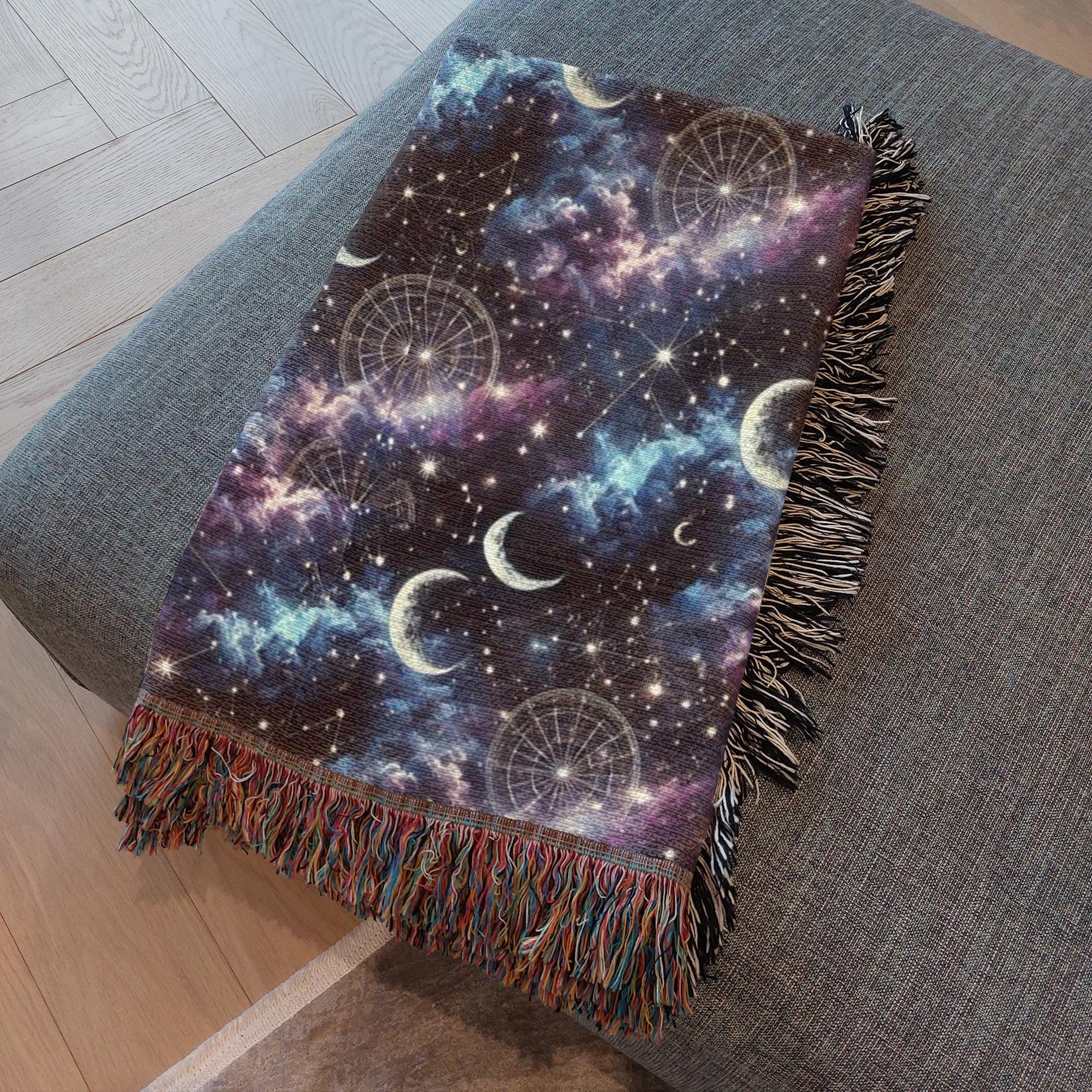 A woven throw blanket with a celestial design featuring crescent moons, constellations, and a dreamy night sky in shades of navy, purple, and blue is neatly draped over the arm of a gray upholstered couch. The blanket has multicolored fringe on one end and black-and-white fringe on the other, adding a cozy and artistic touch to the modern living space with light wood flooring.
