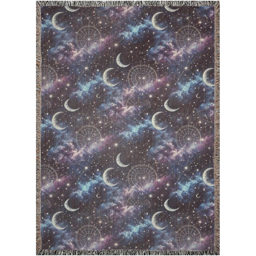 Flat-lay image of a woven tapestry blanket featuring a celestial design with crescent moons, star constellations, and dreamy clouds in shades of purple, blue, and black. The blanket is bordered with multicolored fringe, enhancing its boho and mystical aesthetic.