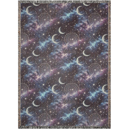 Flat-lay image of a woven tapestry blanket featuring a celestial design with crescent moons, star constellations, and dreamy clouds in shades of purple, blue, and black. The blanket is bordered with multicolored fringe, enhancing its boho and mystical aesthetic.