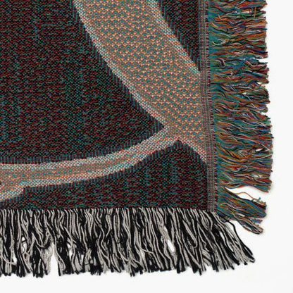 Close-up of the corner of a woven tapestry blanket featuring a multicolor design with curved lines. The blanket has fringed edges in black, white, and multicolored yarns, showcasing the textured weave and detailed craftsmanship.