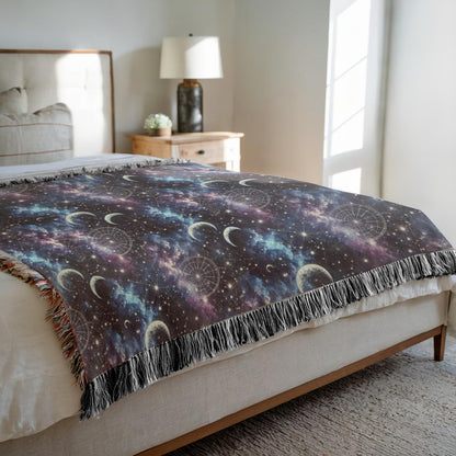 Cozy bedroom scene featuring a woven celestial blanket spread across a neatly made bed. The blanket displays a dreamy night sky design with crescent moons, stars, constellations, and swirling clouds in shades of purple, blue, and black. The edges are finished with multicolored fringe, adding a bohemian touch. A wooden nightstand with a lamp and flower vase sits beside the bed, bathed in soft natural light from a nearby window.