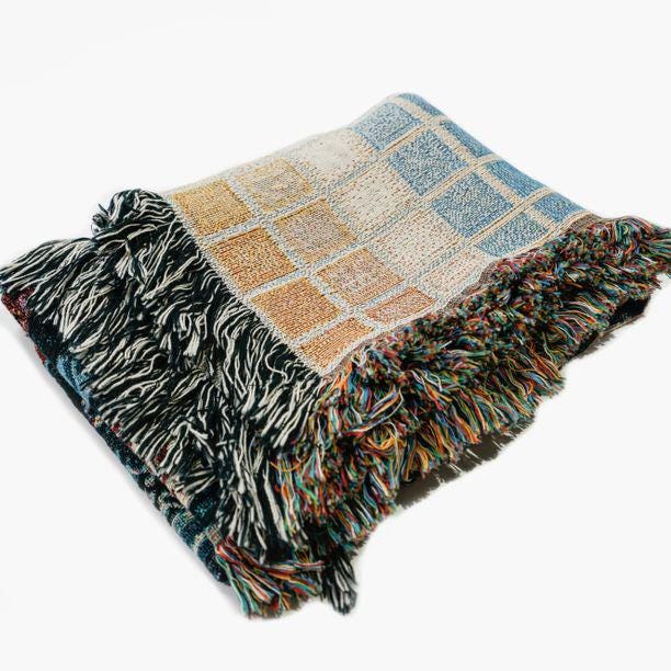 Folded woven blanket featuring a gradient grid pattern with soft pastel squares in shades of blue, peach, and cream. The blanket has multicolored fringe on two sides, showcasing a mix of black, white, and rainbow yarns. The texture and weave are clearly visible against a white background.