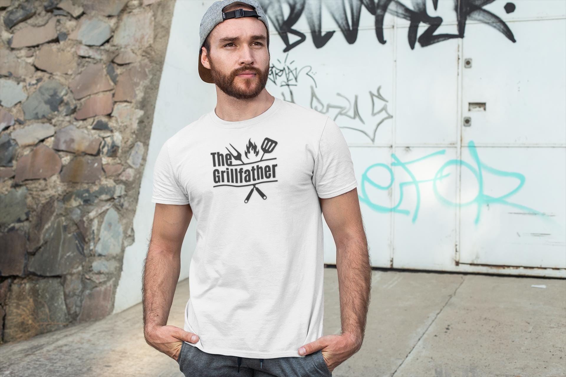 Man standing outdoors against a graffiti-covered wall, wearing a white T-shirt with the phrase “The Grillfather” and grilling icons, along with a gray backward cap.