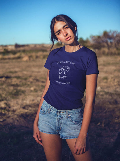 A young woman is standing outdoors in a sunlit field, wearing a navy blue graphic t-shirt and light blue denim shorts. The t-shirt features a white illustration of a rooster&#39;s head with the text &quot;CLUCK AROUND&quot; above and &quot;AND FIND OUT&quot; below. She has long dark hair parted in the middle, and her head is slightly tilted as she gazes at the camera. The background shows dry grass and scattered trees under a clear blue sky.
