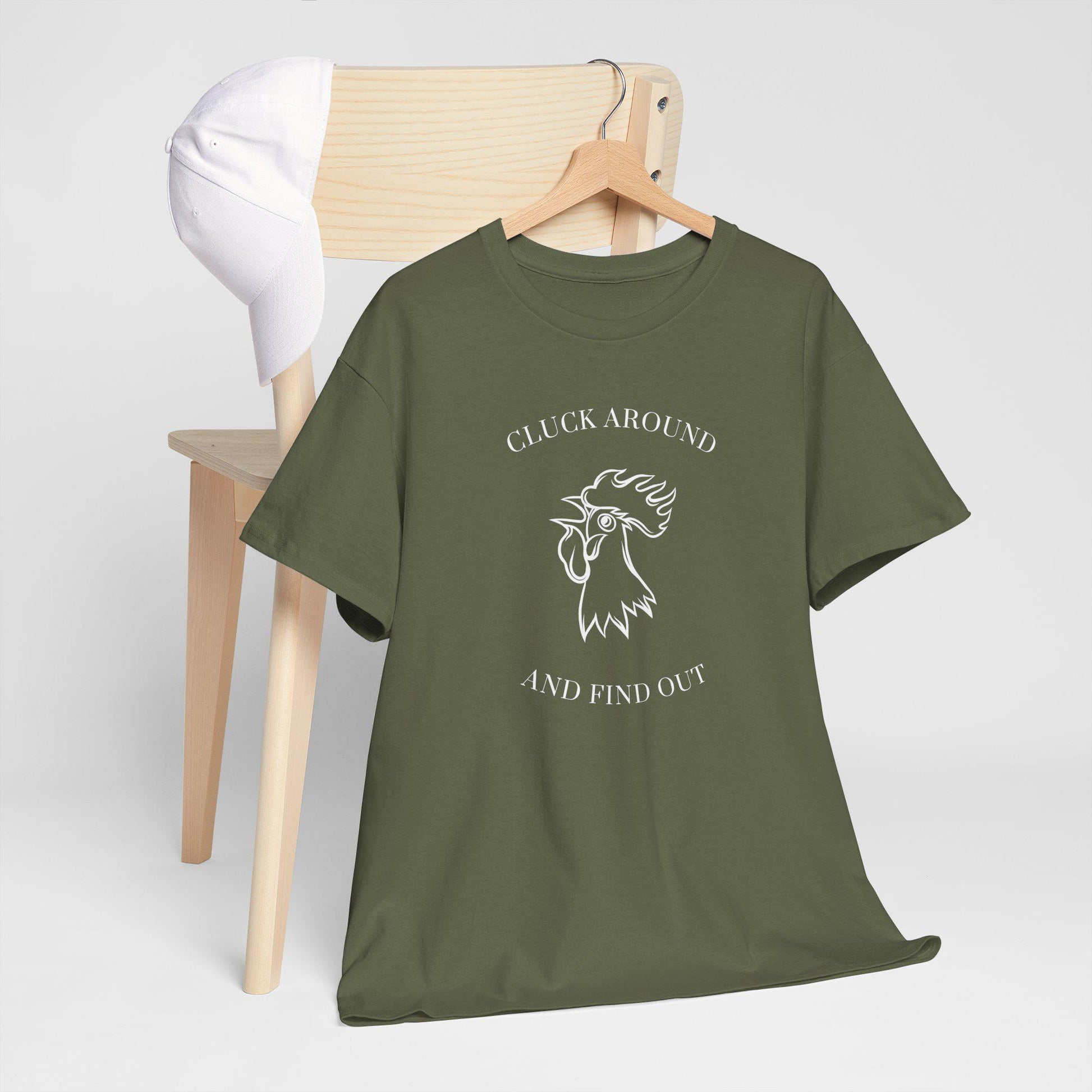 An olive green t-shirt with a white graphic design is draped over a light wooden chair. The design features a stylized rooster head with the text &quot;CLUCK AROUND&quot; above and &quot;AND FIND OUT&quot; below. A white baseball cap rests on the back of the chair. The scene is set against a simple light gray background, giving it a clean and minimal look.