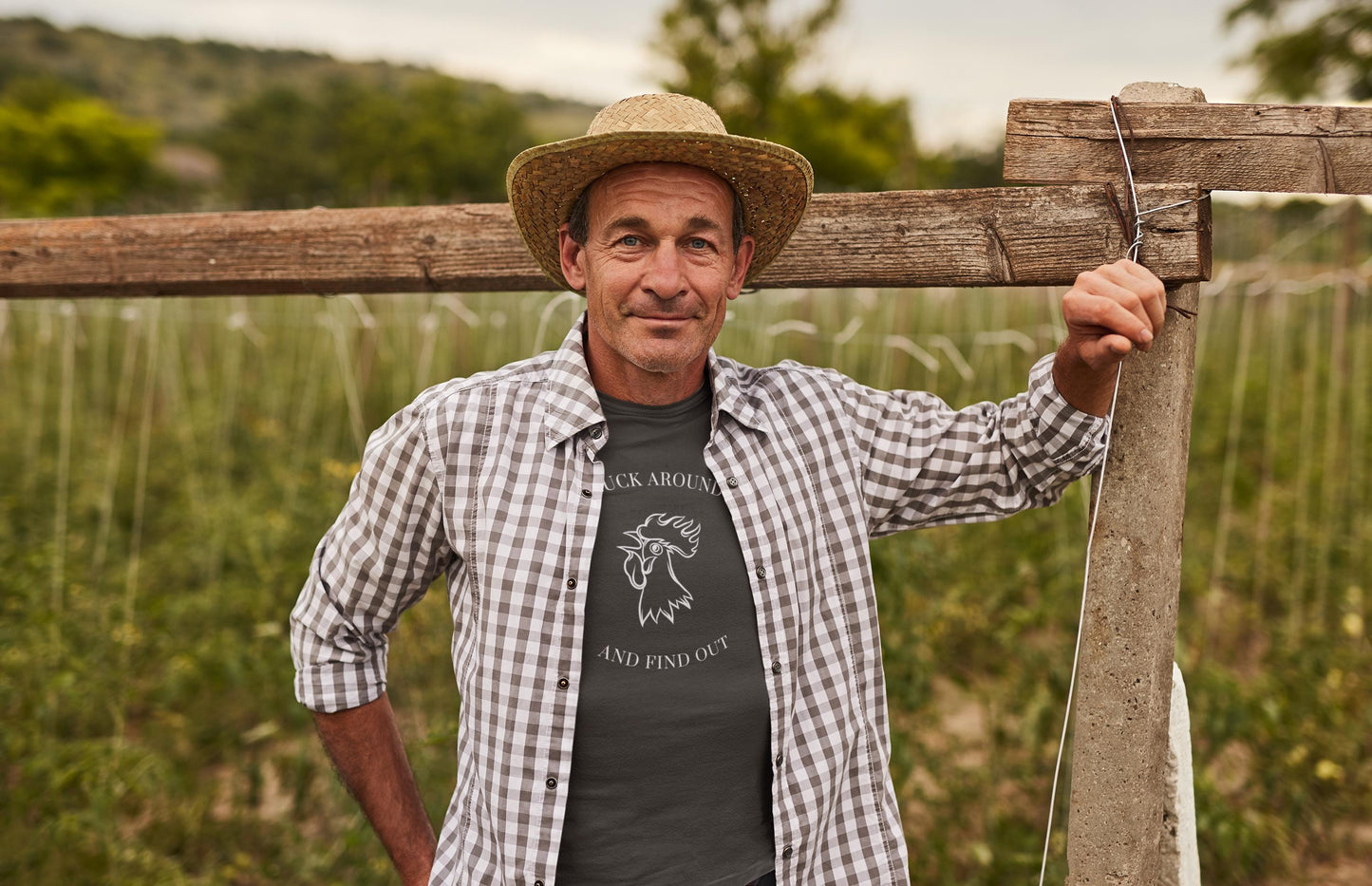 A middle-aged man stands outdoors in a green, rural field, leaning on a wooden fence post. He is wearing a straw hat, a dark graphic t-shirt with a rooster illustration and the text &quot;CLUCK AROUND AND FIND OUT,&quot; and a black-and-white plaid button-up shirt worn open. He has a relaxed and confident expression, with trees and crops visible in the background under a cloudy sky.