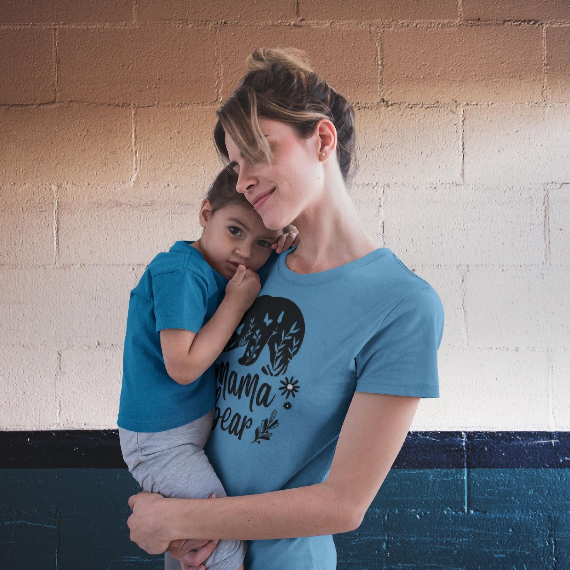 Loving mother holding her young child, both wearing matching blue t-shirts. The mother’s shirt features a graphic of a bear with trees inside it and the text &#39;Mama Bear&#39; surrounded by floral elements. They are standing against a textured wall with beige and dark blue color blocks.