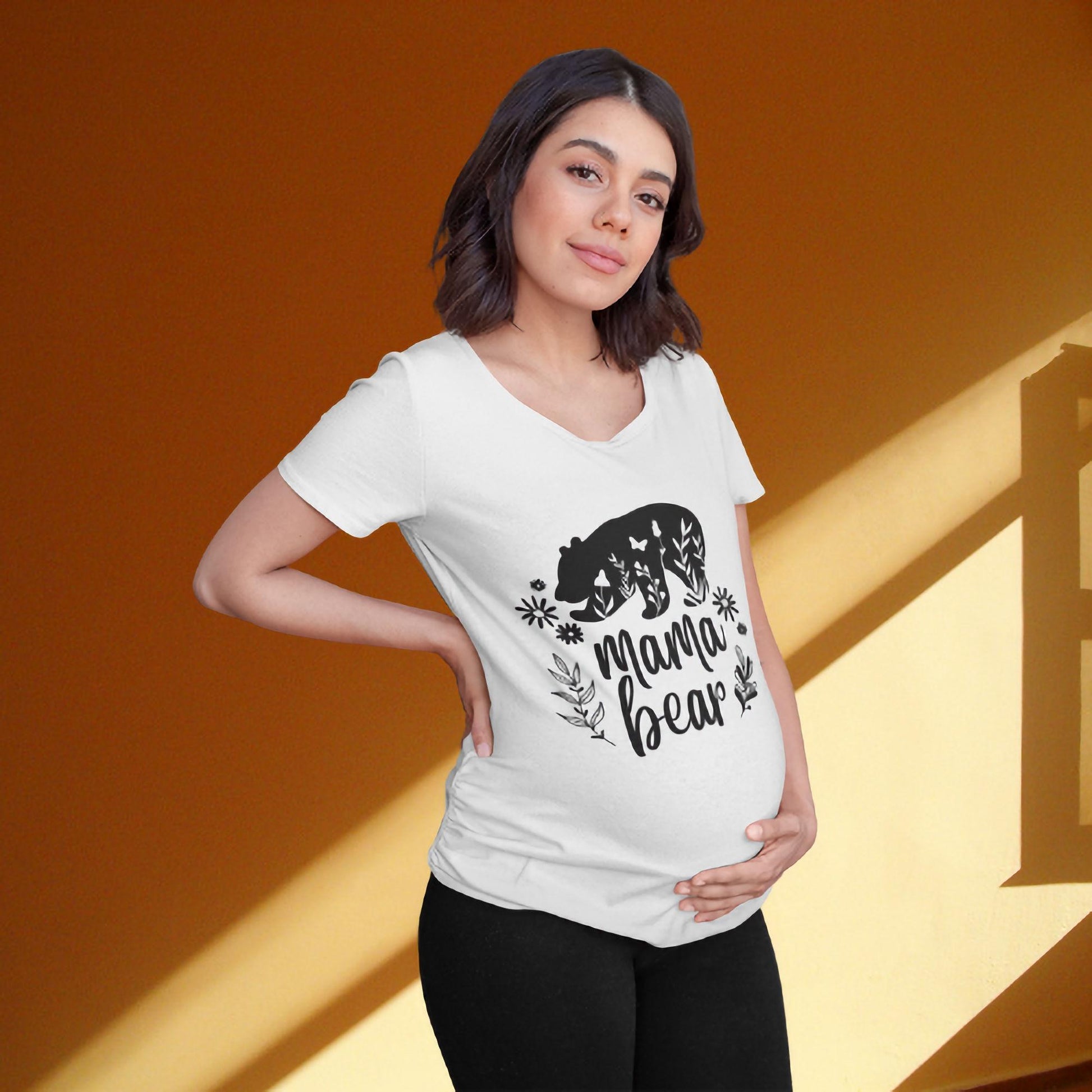 Pregnant woman gently cradling her belly while wearing a white maternity t-shirt with a black graphic of a bear, floral accents, and the phrase &#39;Mama Bear.&#39; She stands against a warm orange wall with sunlight streaming through a window, creating a cozy and serene atmosphere.
