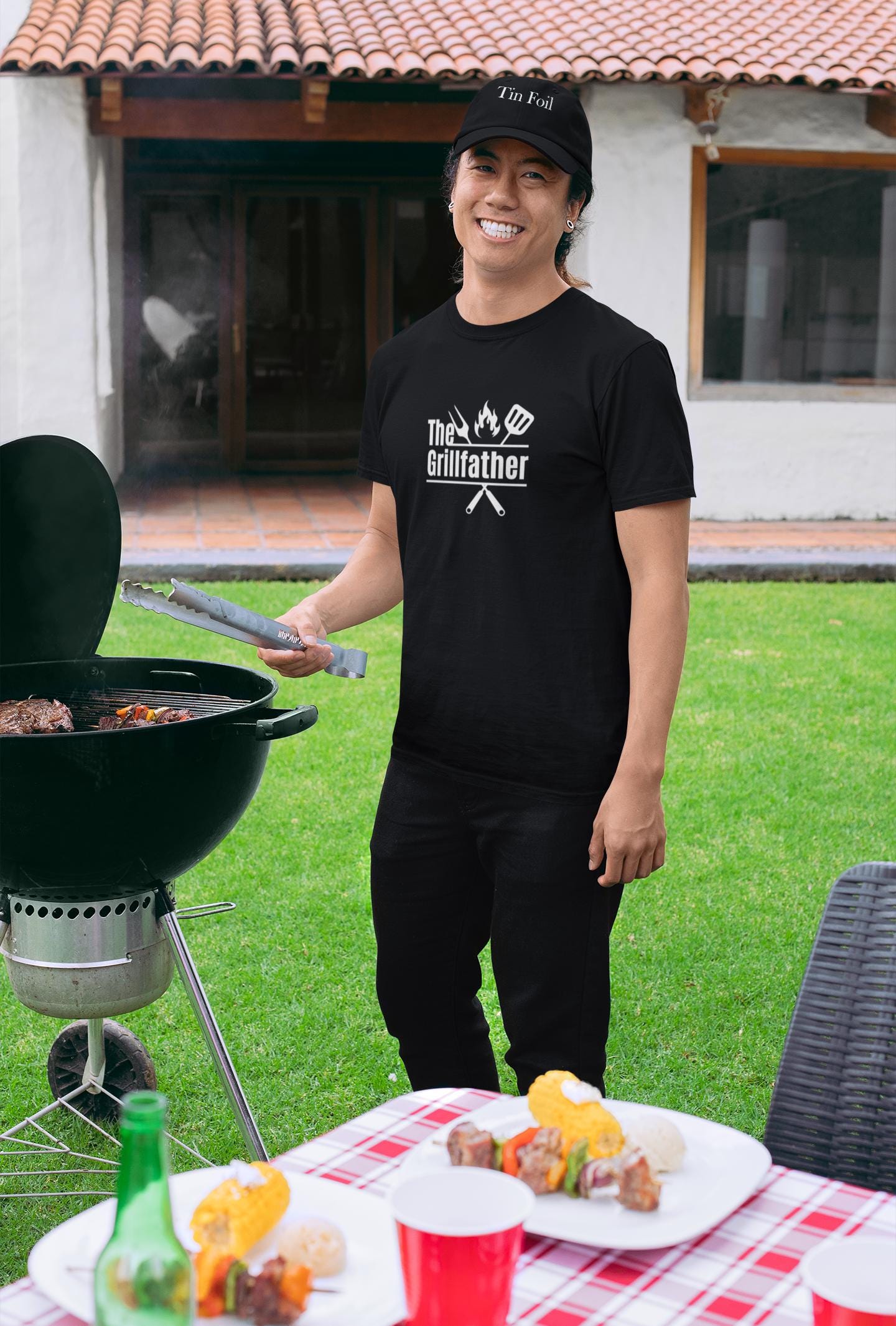 Smiling man grilling outdoors, wearing a black T-shirt with the phrase “The Grillfather” and grilling icons, along with a black cap. A picnic table with food and drinks is set in front of him.