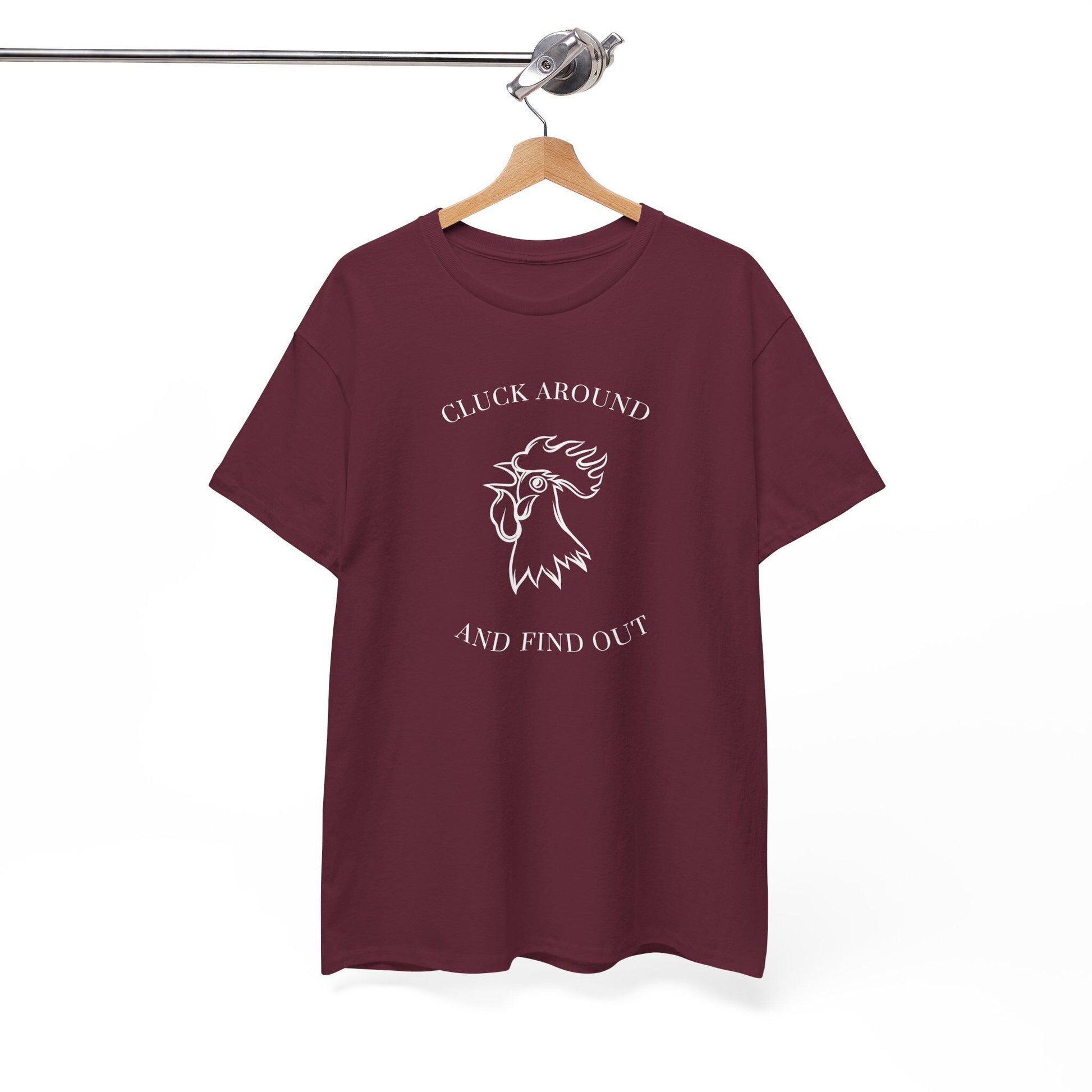A maroon t-shirt is displayed on a wooden hanger suspended from a metal clothing rack. The shirt features a white graphic design of a rooster&#39;s head with the text &quot;CLUCK AROUND&quot; above and &quot;AND FIND OUT&quot; below. The shirt has a classic crew neckline and relaxed fit, hanging against a plain white background.