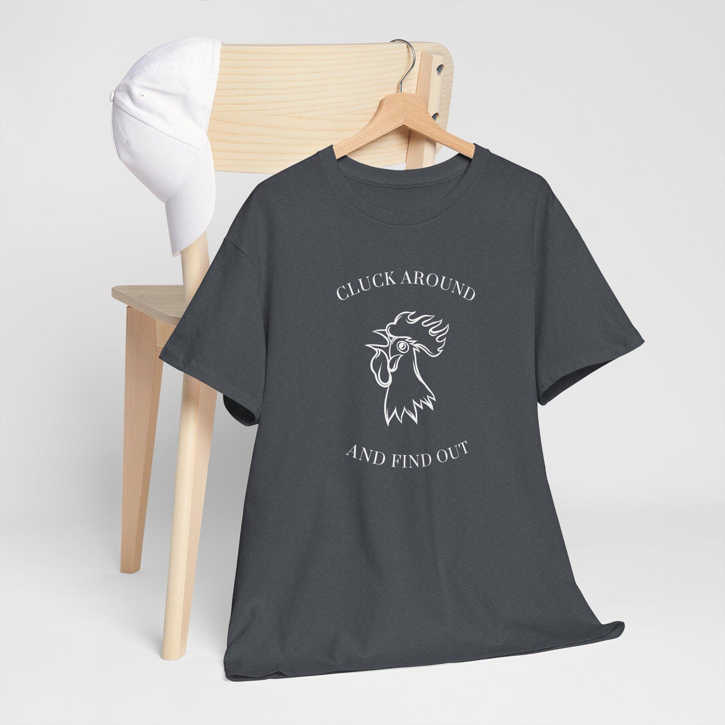 A dark gray t-shirt with a white graphic design is draped over a wooden chair. The design features an illustrated rooster head with the text &quot;CLUCK AROUND&quot; above and &quot;AND FIND OUT&quot; below. A white baseball cap is resting on the backrest of the chair. The setup is placed against a plain light gray background, giving a clean and minimalistic look.