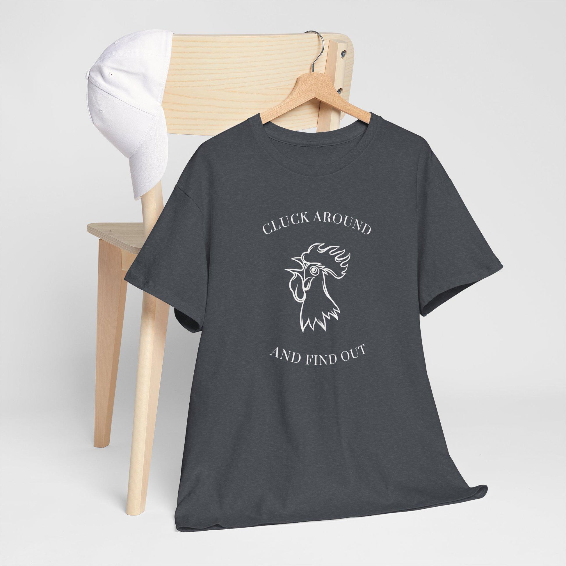 A dark gray t-shirt with a white graphic design is draped over a wooden chair. The design features an illustrated rooster head with the text &quot;CLUCK AROUND&quot; above and &quot;AND FIND OUT&quot; below. A white baseball cap is resting on the backrest of the chair. The setup is placed against a plain light gray background, giving a clean and minimalistic look.