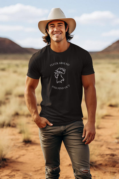 A young man is standing in a desert landscape with mountains in the background, smiling confidently. He is wearing a light beige cowboy hat, a black graphic t-shirt with a white outline of a rooster and the text &quot;CLUCK AROUND AND FIND OUT,&quot; and distressed gray jeans. The sky is partly cloudy, and the natural lighting highlights the rugged and western-inspired vibe of the scene.