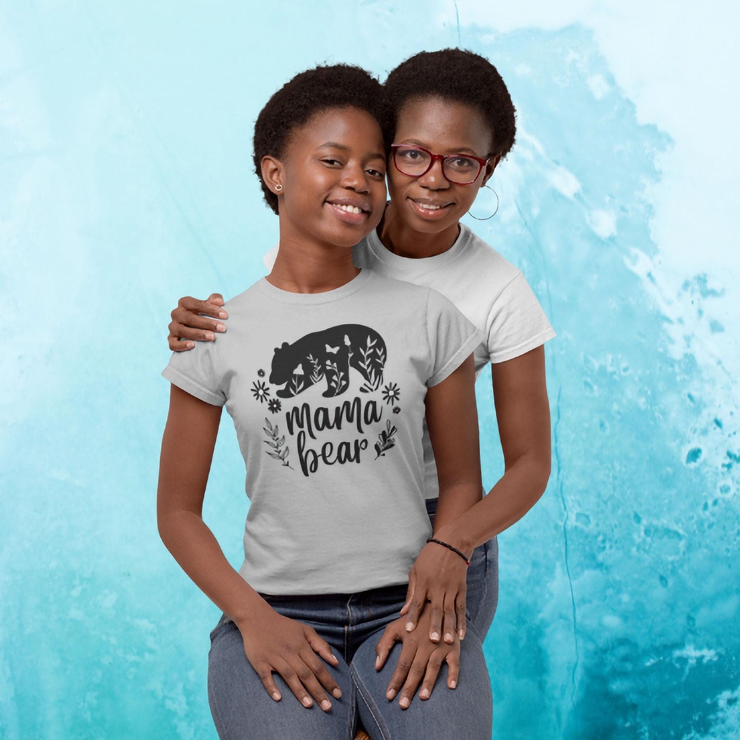 Smiling mother and daughter posing together in front of a light blue textured background. The daughter is wearing a light gray t-shirt featuring a black bear graphic, floral elements, and the text &#39;Mama Bear.&#39; The mother, standing behind and hugging her daughter, wears a plain white t-shirt and red glasses