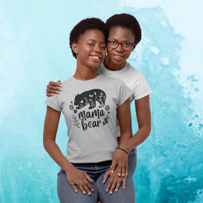 Smiling mother and daughter posing together in front of a light blue textured background. The daughter is wearing a light gray t-shirt featuring a black bear graphic, floral elements, and the text &#39;Mama Bear.&#39; The mother, standing behind and hugging her daughter, wears a plain white t-shirt and red glasses