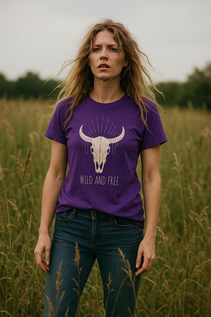 Woman standing in a grassy field wearing a purple T-shirt with a graphic of a bull skull and radiating decorative lines above it. The shirt also features the text “WILD AND FREE” below the design. She has long wavy hair, wears blue jeans, and has a serious expression.