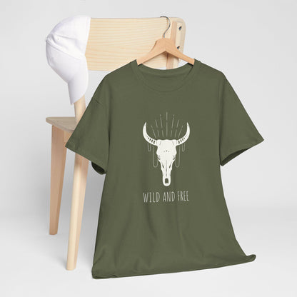 Olive green T-shirt displayed on a wooden hanger, draped over the back of a light wooden chair with a white cap hanging on the corner. The shirt features a graphic of a white bull skull with decorative lines above it and the text “WILD AND FREE” below. Shown against a plain light background.