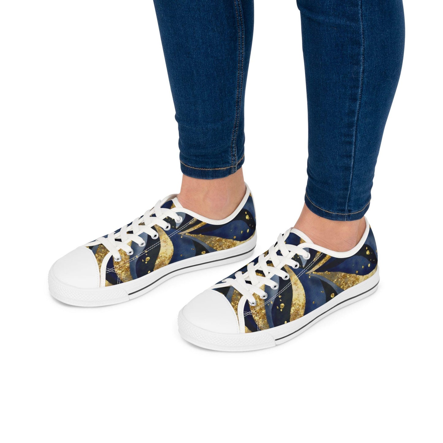 A person wearing low-top canvas sneakers with a cosmic-themed design in deep blue and gold tones. The shoes feature white soles, white laces, and white toe caps. The wearer is dressed in fitted blue jeans, and the background is a clean white, drawing attention to the stylish, celestial-patterned footwear.