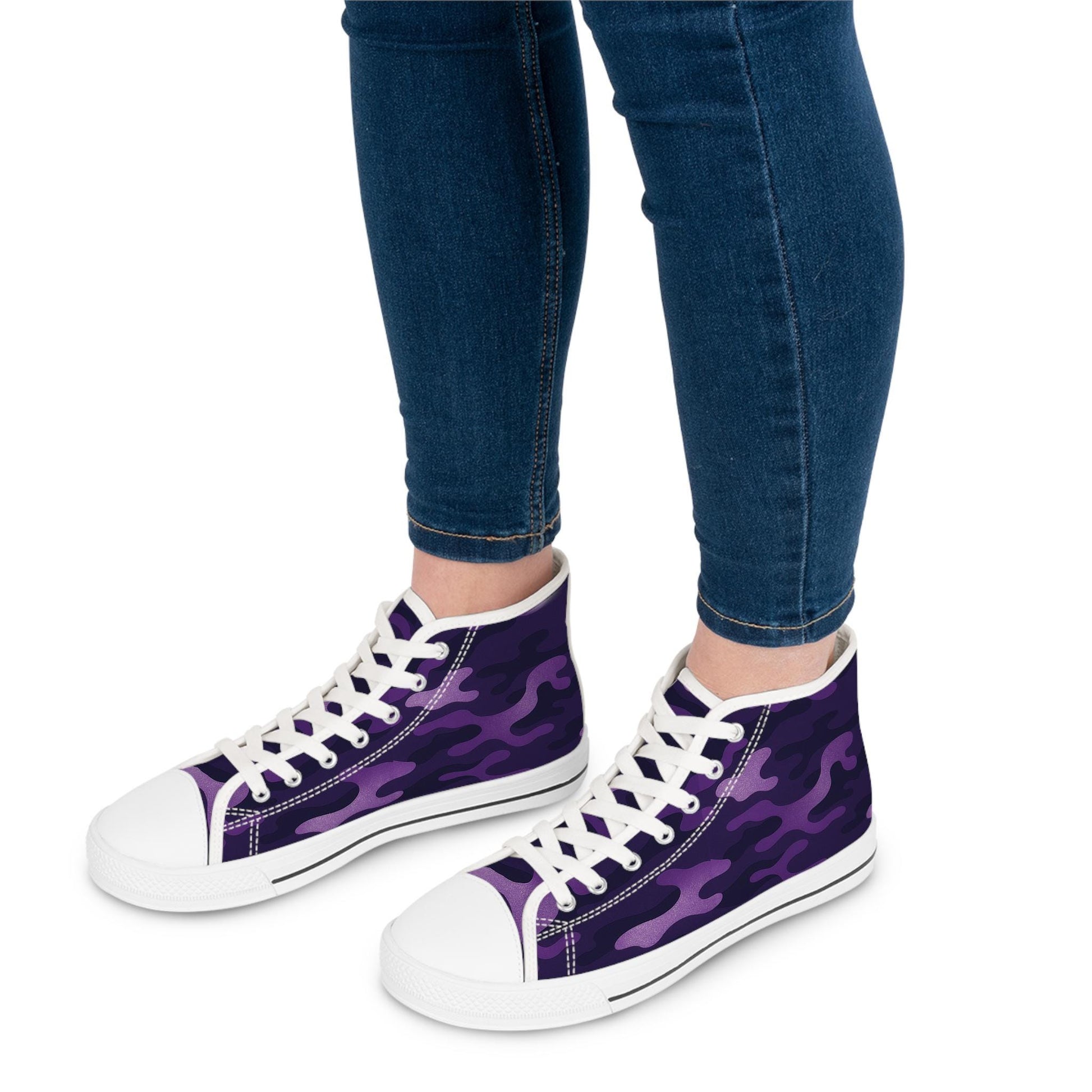 A person wearing dark blue skinny jeans is shown from the calves down, modeling a pair of high-top sneakers with a vibrant purple camouflage pattern. The shoes feature white rubber toe caps, white soles with a black stripe, and white laces, creating a bold and stylish contrast.