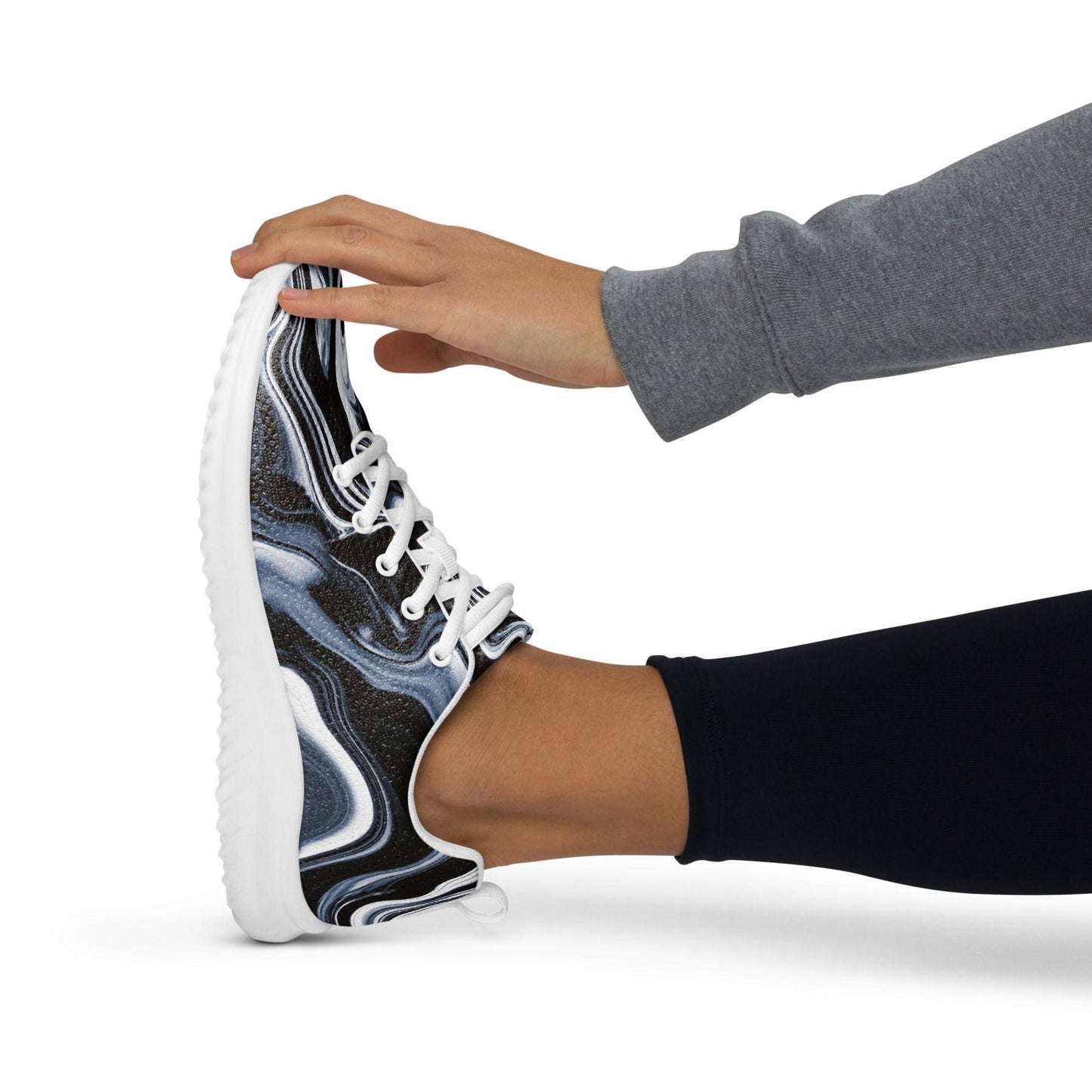 Woman stretching while wearing stylish black, white, and gray swirl-patterned athletic sneakers with white laces and cushioned soles. Modern activewear shoe ideal for fitness routines, yoga, running, or casual workouts—designed for comfort, flexibility, and bold artistic flair.