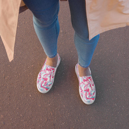 A top-down view of a person standing on a paved surface, wearing light blue skinny jeans and slip-on sneakers with a vibrant pink, white, and teal marbled swirl pattern. The person is also wearing a beige or light tan coat, partially visible in the frame.