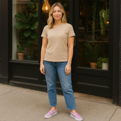 A woman stands on a city sidewalk in front of a storefront with large windows and potted plants inside. She is wearing a beige t-shirt, blue jeans, and slip-on sneakers featuring a pink, white, and blue marbled swirl pattern. She is smiling slightly and looking at the camera in a relaxed, casual pose.