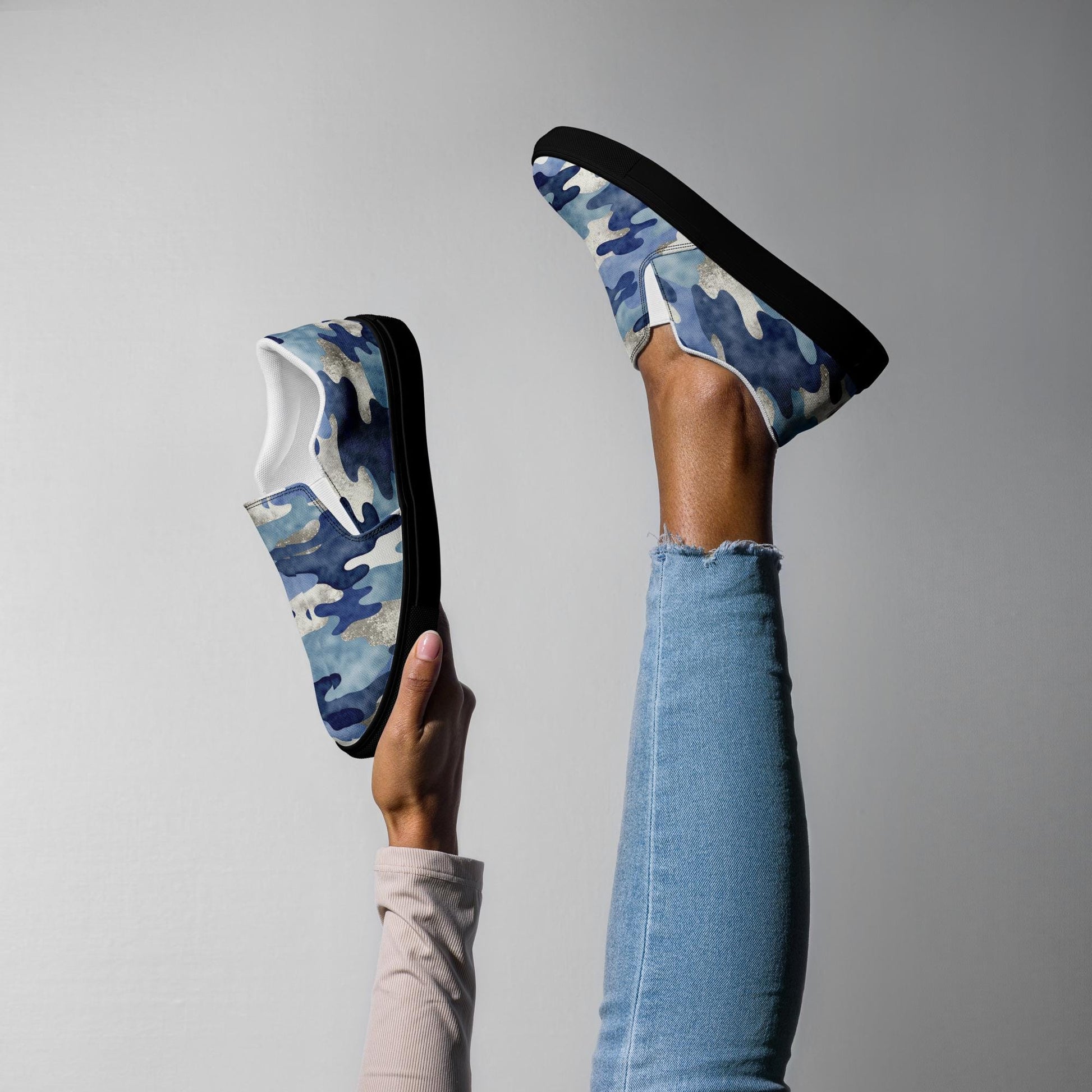 Women’s blue camo slip-on canvas sneakers with black soles, modeled in playful pose with leg raised and hand holding shoe, trendy casual footwear.