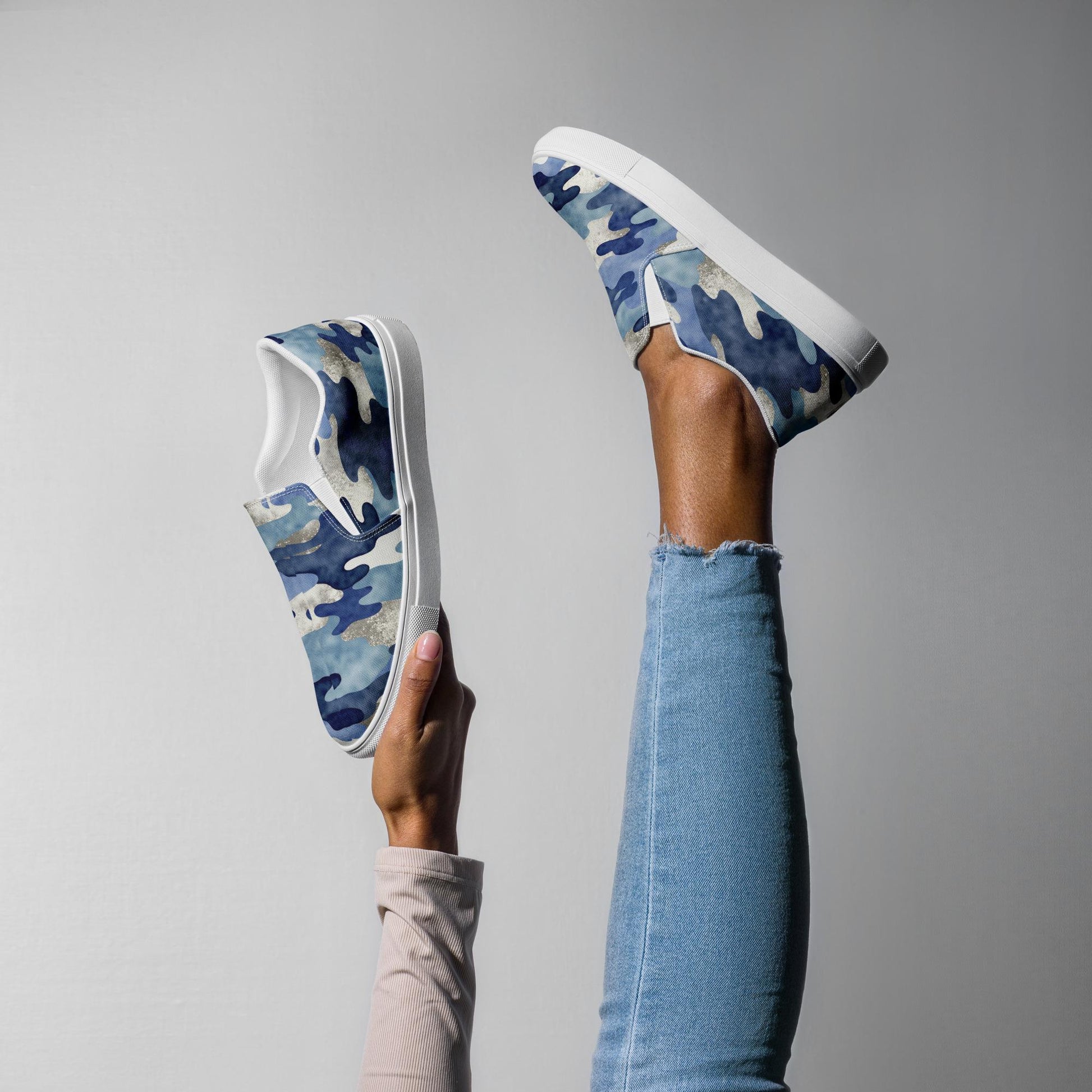 Women’s blue camo slip-on canvas shoes with white soles, styled in playful pose with leg raised and hand holding shoe, casual and comfortable footwear.
