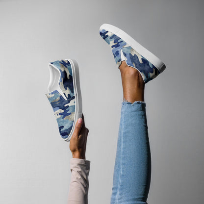 Women’s blue camo slip-on canvas shoes with white soles, styled in playful pose with leg raised and hand holding shoe, casual and comfortable footwear.