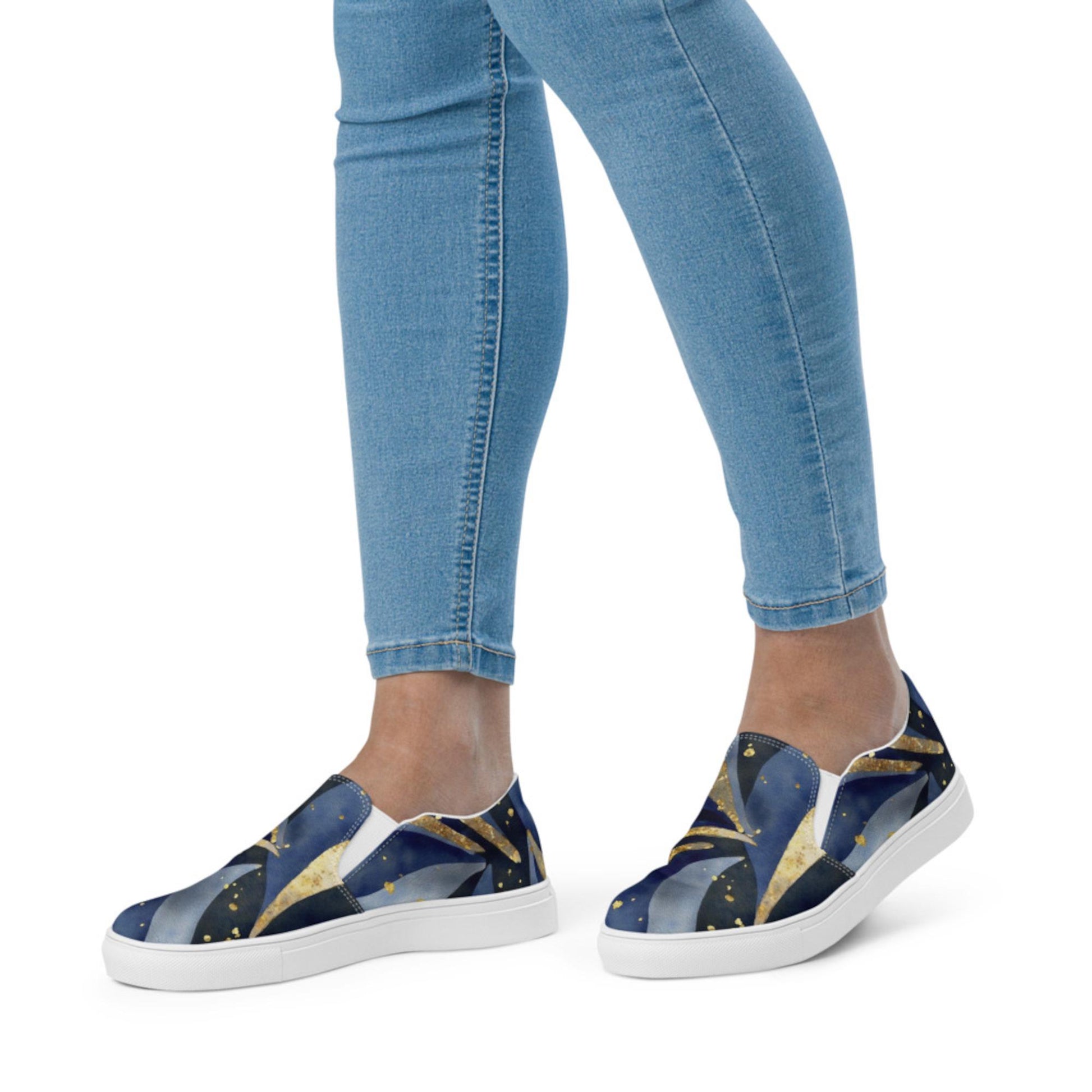 A person wearing slip-on sneakers with a celestial-themed design featuring swirling deep blue, black, and gold patterns resembling stars and cosmic waves. The shoes have clean white soles and a low-profile silhouette. The individual is dressed in light blue skinny jeans, and the background is plain white, emphasizing the stylish and artistic footwear.