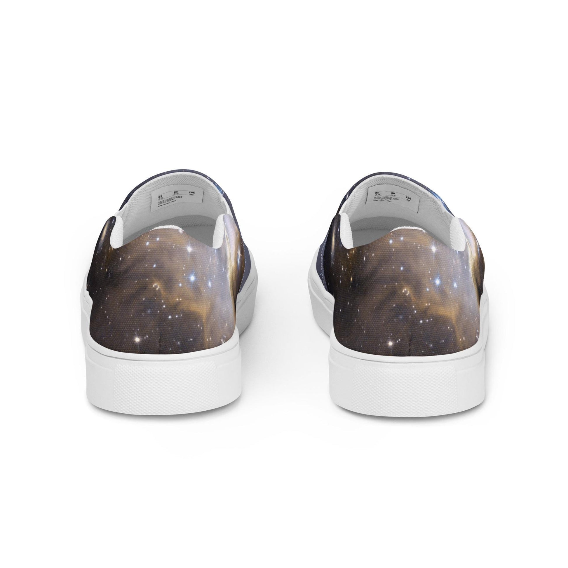 Rear view of a pair of slip-on sneakers featuring a galaxy-themed design with dark cosmic clouds and scattered stars in gold, blue, and black tones. The shoes have white inner linings and white rubber soles, displayed against a clean white background.