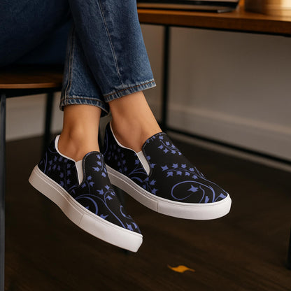 Close-up of a person wearing black slip-on sneakers with a deep purple ivy vine pattern, sitting indoors on a wooden chair. The shoes feature white soles and are paired with rolled-up blue jeans, creating a comfortable and stylish casual look.