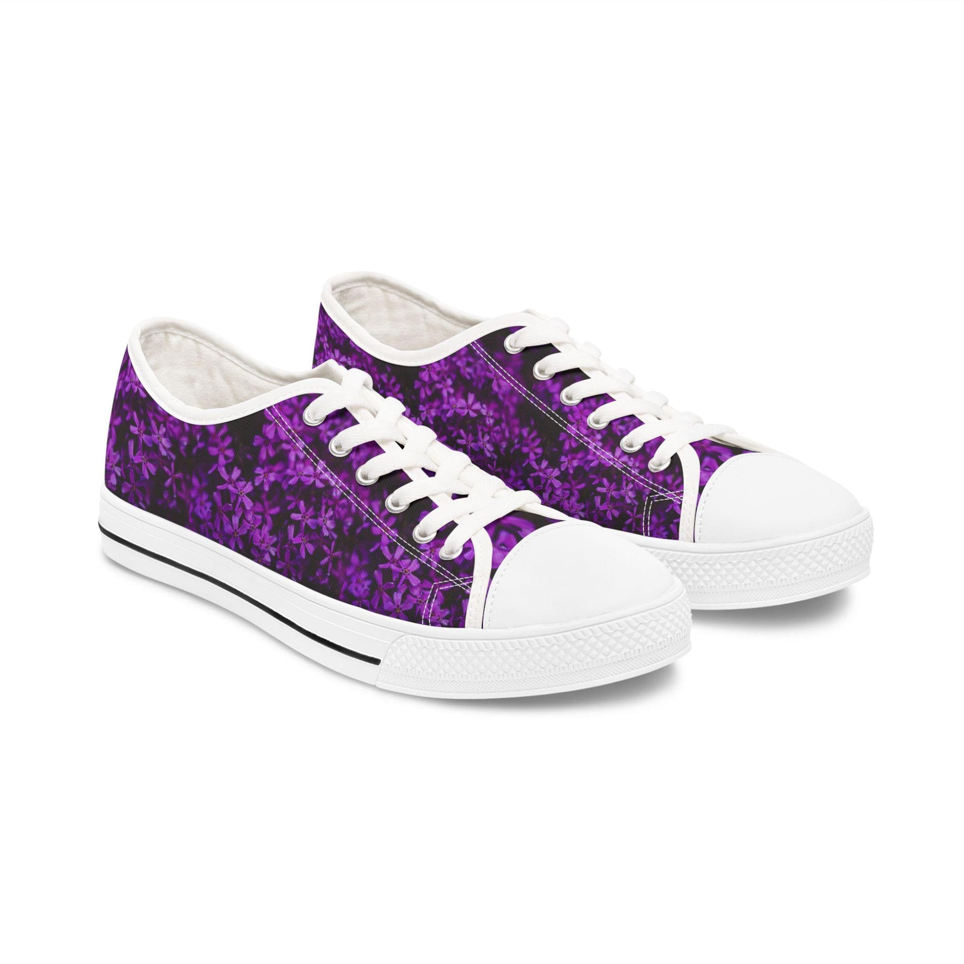 A pair of low-top sneakers is displayed on a white background. The shoes feature a vibrant purple floral pattern on a dark background. They have white laces, white rubber toe caps, and white soles with a black stripe, creating a crisp and stylish contrast with the bold floral design.