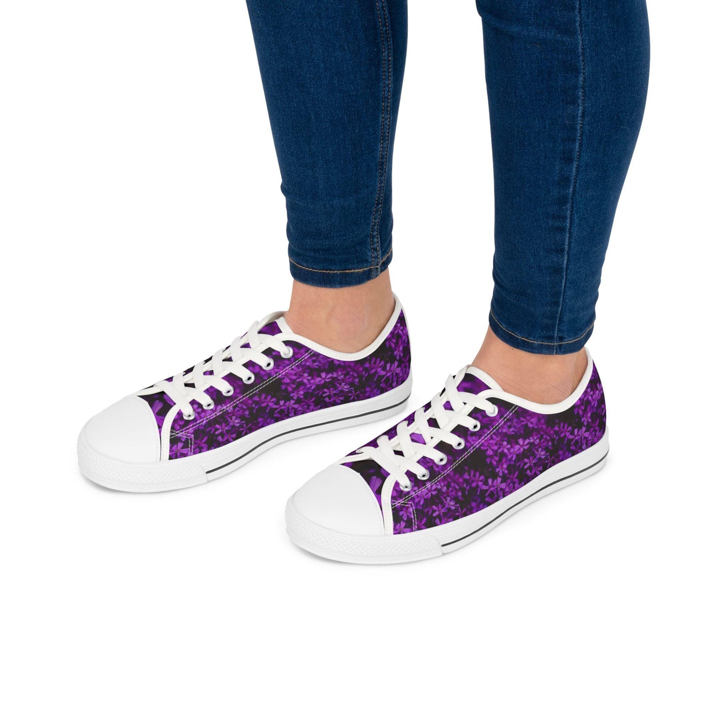 A person wearing dark blue skinny jeans is modeling a pair of low-top sneakers with a bold purple floral pattern on a dark background. The shoes feature white laces, white rubber toe caps, and white soles with a thin black stripe, creating a stylish contrast with the vibrant upper design. The background is plain white, highlighting the footwear.