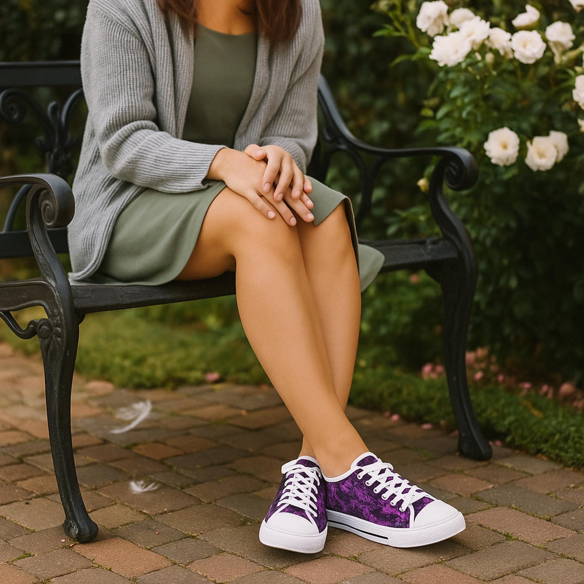A woman is seated on a decorative black iron bench in a garden with white roses in the background. She is wearing a sage green dress paired with a light gray cardigan. On her feet are low-top sneakers with a rich purple floral pattern, white laces, white rubber toe caps, and white soles with a black stripe. The setting is peaceful and charming, with a cobblestone path and greenery surrounding the bench.
