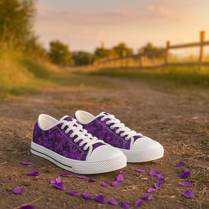 A pair of low-top sneakers with a rich purple floral pattern, white laces, white rubber toe caps, and white soles is placed on a dirt path at sunset. Scattered purple flower petals surround the shoes. The background features a wooden fence, grassy fields, and trees bathed in warm golden light, creating a peaceful and romantic outdoor setting.