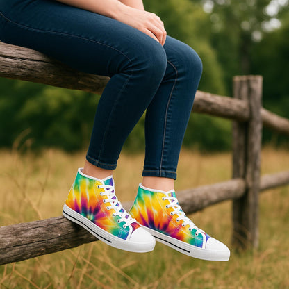 A person wearing dark skinny jeans is sitting on a wooden fence in a grassy field. They are wearing vibrant high-top sneakers with a colorful tie-dye pattern in shades of orange, yellow, green, blue, and purple. The shoes feature white laces, white rubber toe caps, and white soles with a black stripe. The background includes tall grass and blurred green trees, suggesting a peaceful outdoor setting.